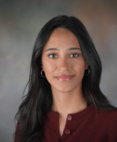 A young woman with long dark hair wearing silver hoop earrings and a burgundy top, smiling slightly, posed against a neutral gray background—perfect for a staff photo at a Mission Valley preschool in San Diego.