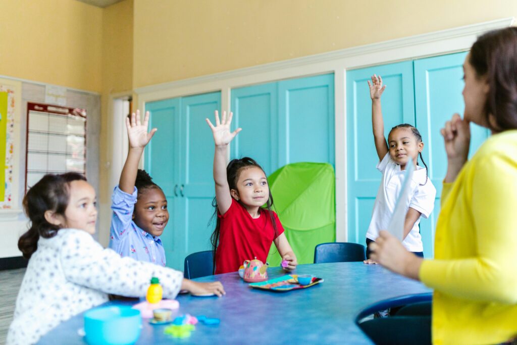 Four young children sit at a round table in a classroom, eagerly raising their hands toward a teacher who engages the group. Classroom supplies are visible on the table, reflecting the importance of early education and child success in preschool programs.