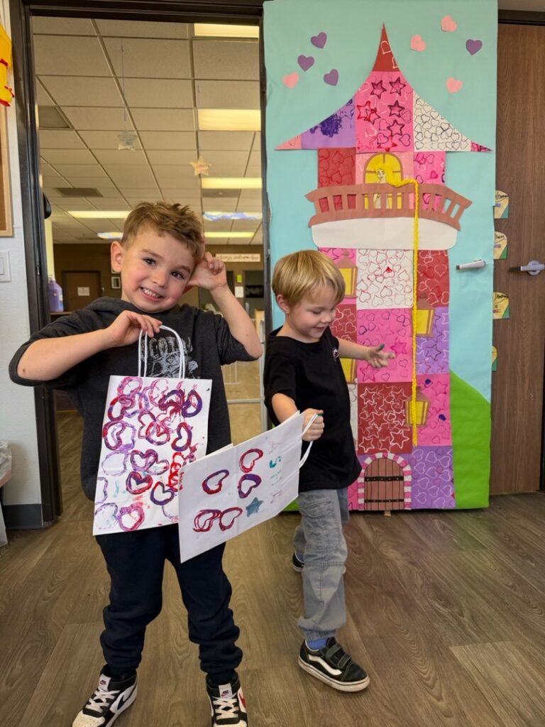 Two young boys in a classroom stand in front of a colorful paper castle mural. One boy smiles and holds decorated bags, while the other looks down and walks away. The scene is cheerful and playful.
