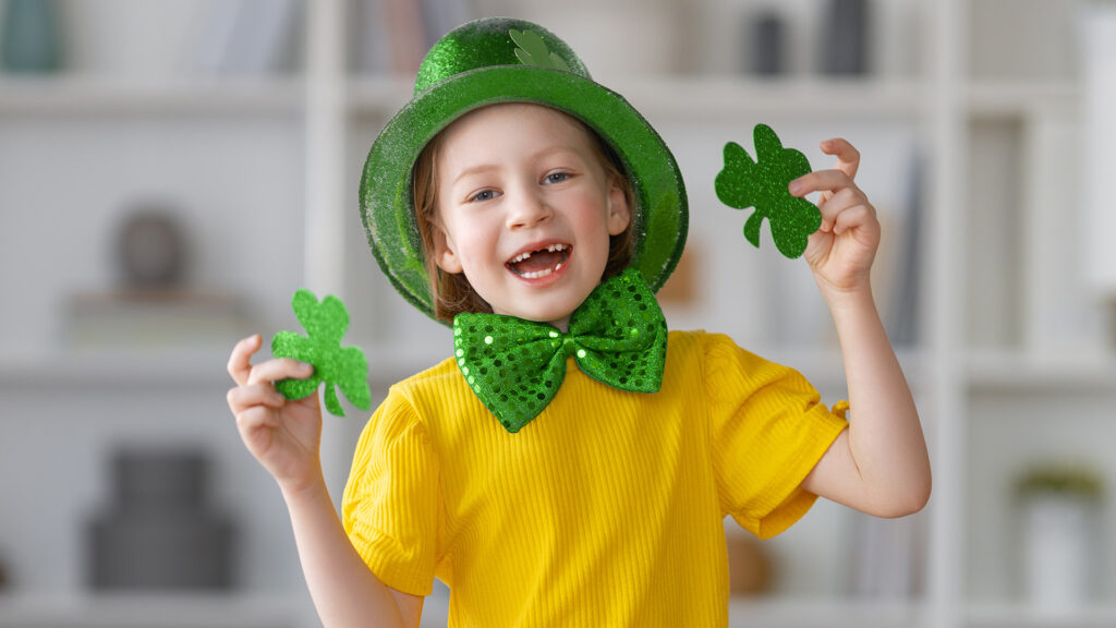 Smiling child wearing a sparkly green hat and bow tie, holding two glittery shamrocks, dressed in a yellow shirt, celebrating St. Patricks Day indoors.