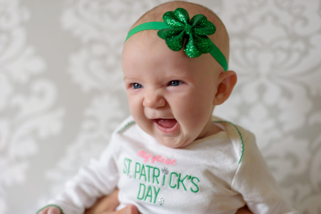 A smiling baby wearing a white shirt that says My first St. Patricks Day and a green headband with a glittery shamrock, held up by an adult in front of a light, patterned background.