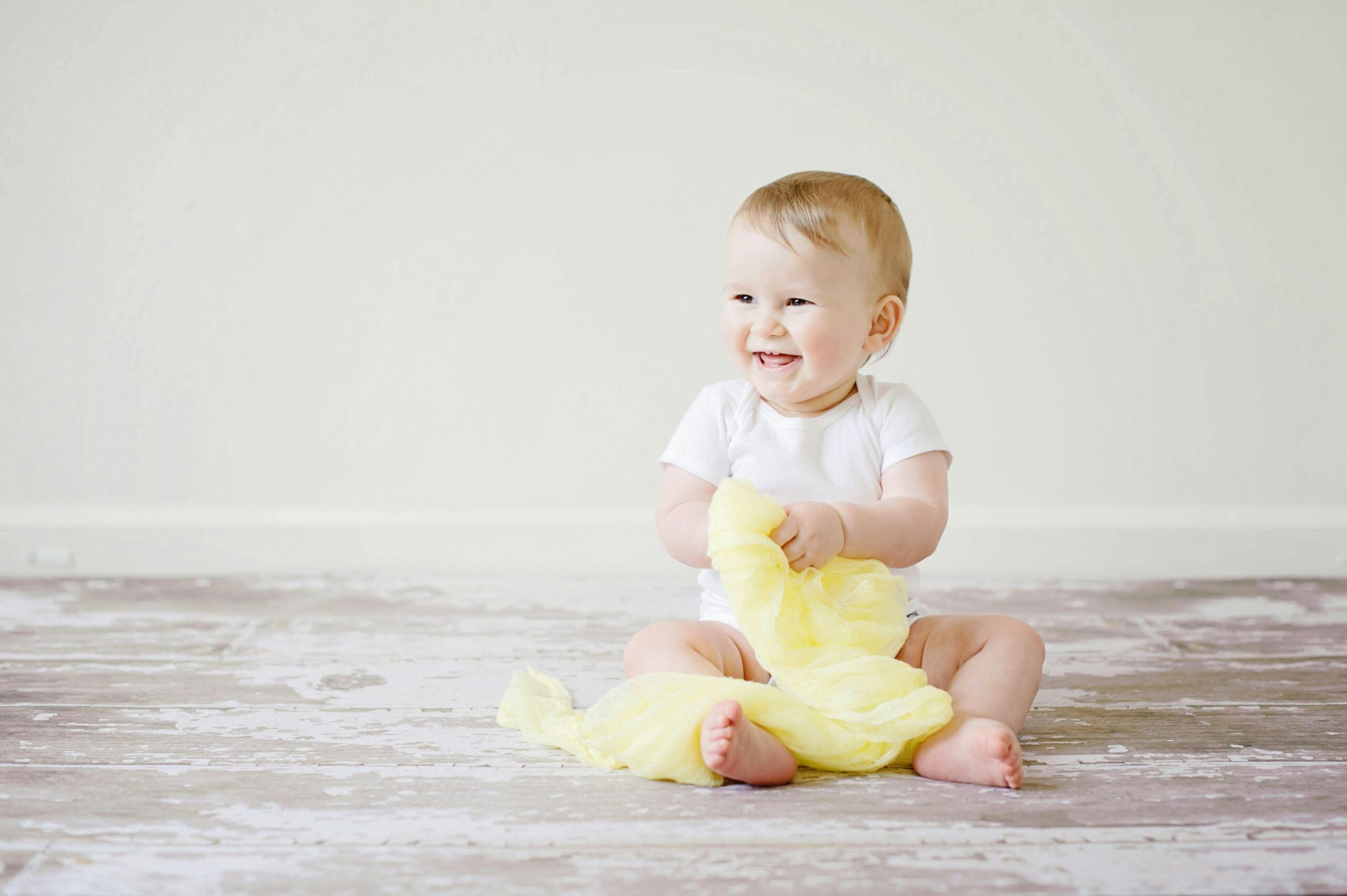 A smiling baby in a white onesie sits on a wooden floor holding a soft yellow cloth, with a light-colored wall in the background.