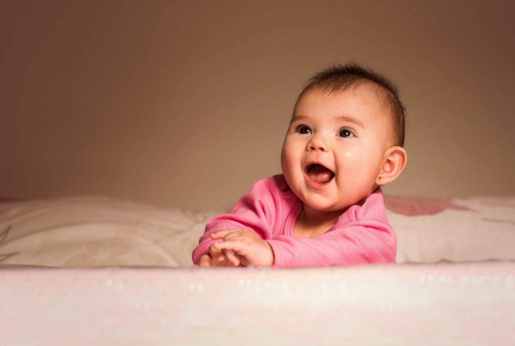 A smiling baby wearing a pink outfit leans forward with hands clasped, looking slightly to the side against a soft, neutral background.