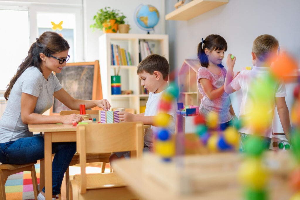 A teacher and a young boy sit at a table playing with colorful blocks in the Ramona preschool program, while two other children play near a chalkboard in a bright, cheerful classroom with books and toys on shelves.
