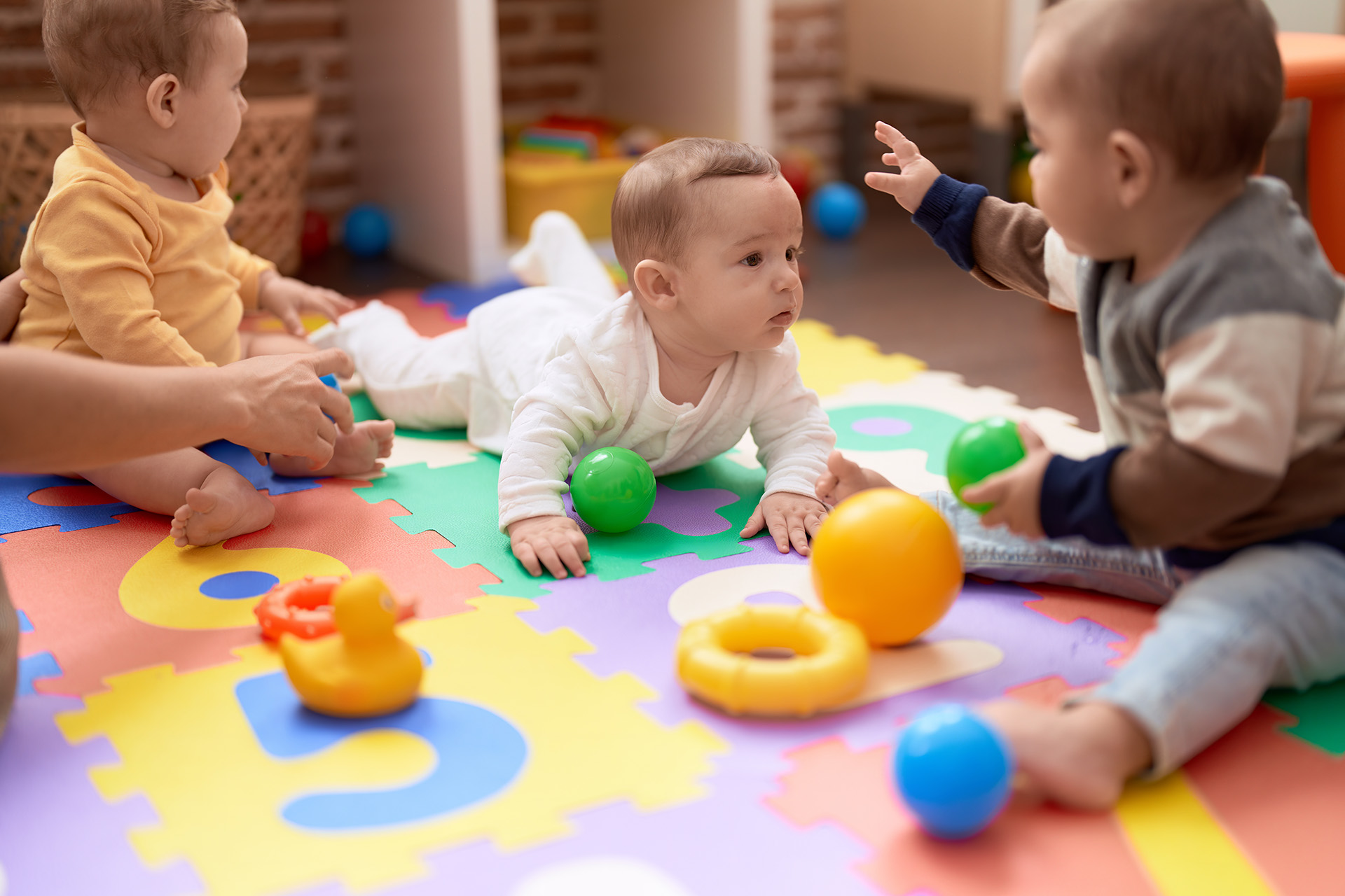 Three babies sit and crawl on colorful foam number mats, playing with various toys including balls and a rubber duck in a bright indoor play area designed for toddler care in Mission Valley, San Diego