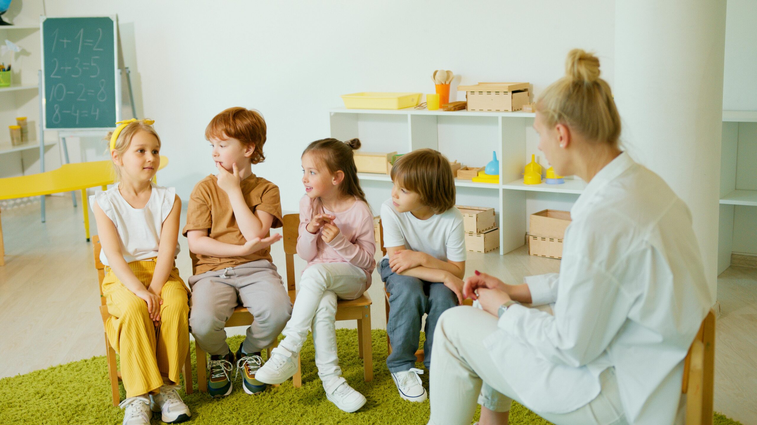 Four young children sit on small chairs in a classroom, talking and smiling, while an adult teacher sits nearby listening. Shelves with educational materials and a chalkboard with math problems are in the background.