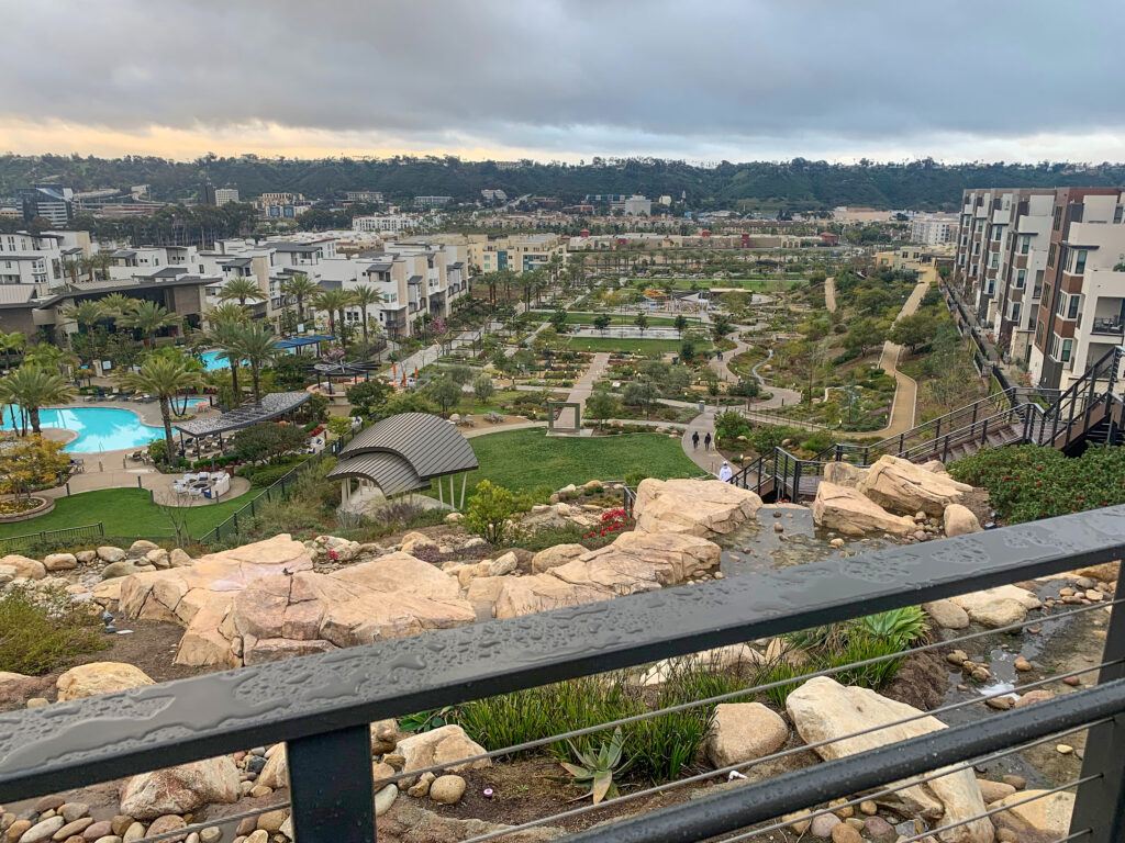 A view from a high terrace overlooking landscaped gardens, walking paths, pools, palm trees, and modern apartment buildings on a cloudy day. Wet railings and rocks are visible in the foreground.
