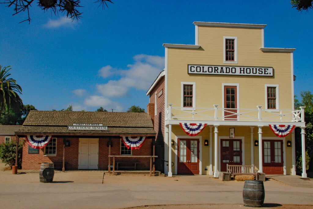 A historic Western-style building labeled Colorado House is decorated with red, white, and blue bunting. Next to it is a brick building labeled Wells Fargo Museum & Store. Both buildings are set under a blue sky in Old Town, San Diego.