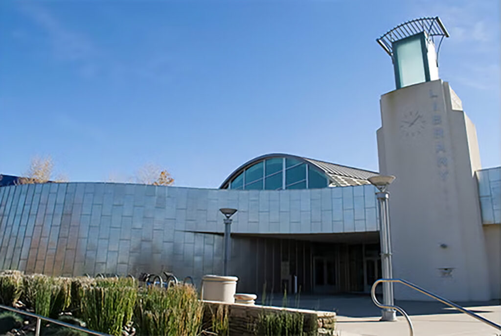 A modern building with a curved metal facade, large windows, a clock tower, and abstract architectural features under a clear blue sky. Steps and landscaped plants are in the foreground.