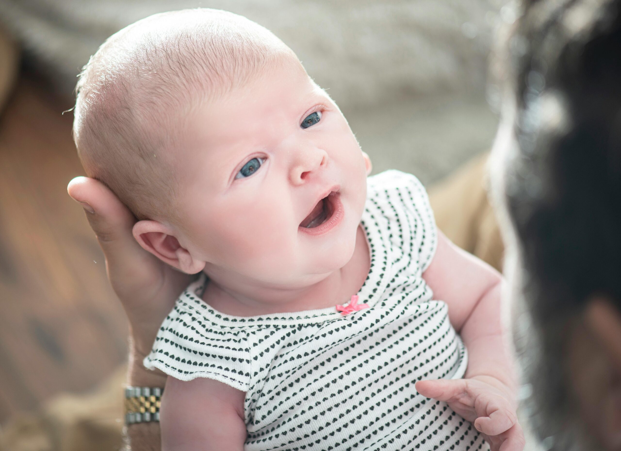 A close-up of a baby with blue eyes and light skin, wearing a white and black striped onesie, being gently held and looking up at an adult whose face is partially visible.