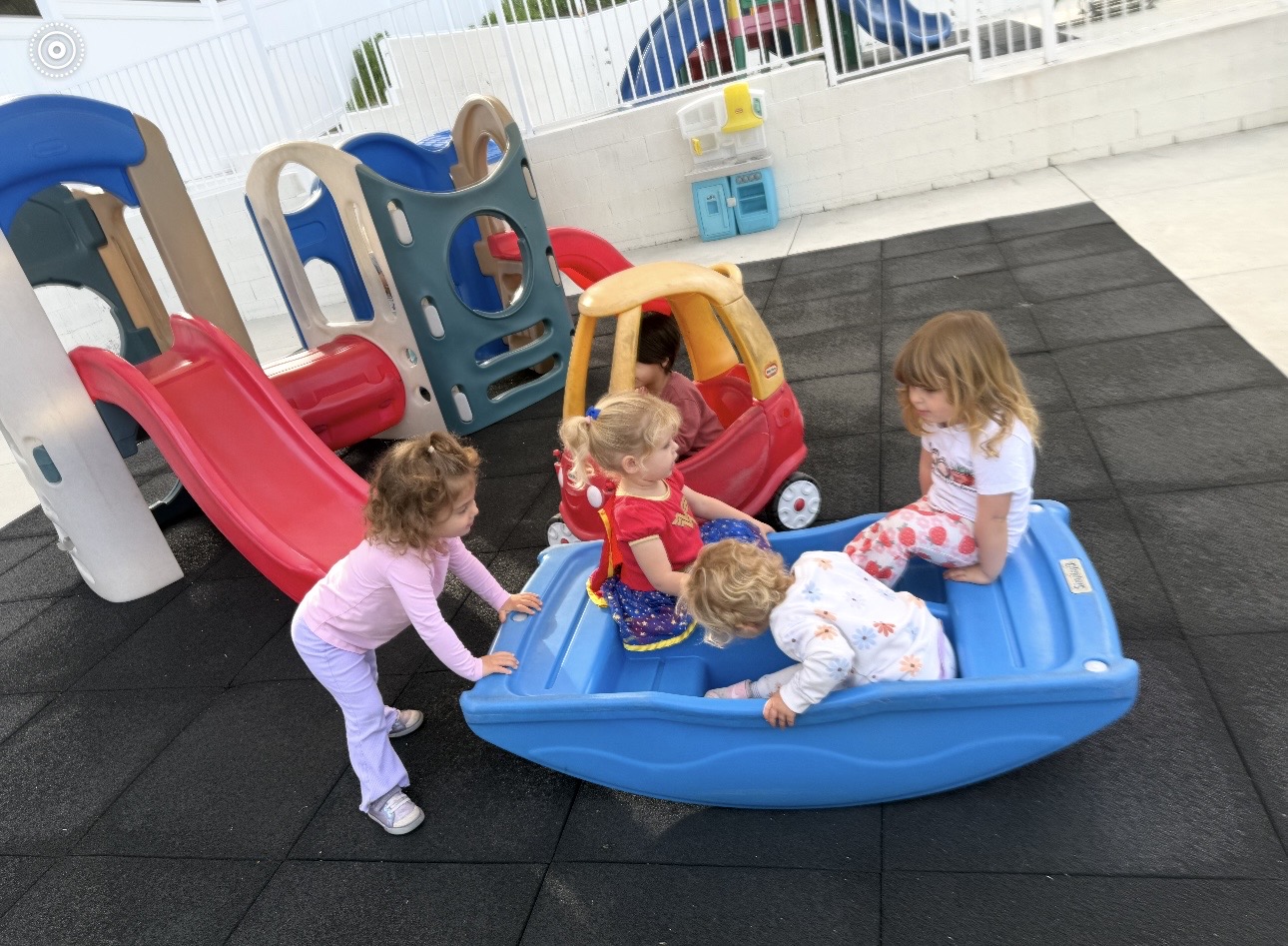 Four young children play together outdoors on playground equipment, including a blue plastic seesaw, a red slide, and a toy car, on a rubber mat surface. They appear to be having fun and interacting with each other.