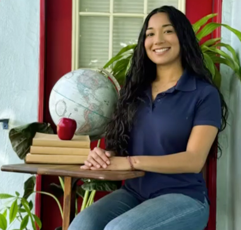 A woman with long dark hair, wearing a navy polo shirt and jeans, sits at a small table with a globe, stacked books, and a red apple in front of a red door and large green plants. She is smiling at the camera.