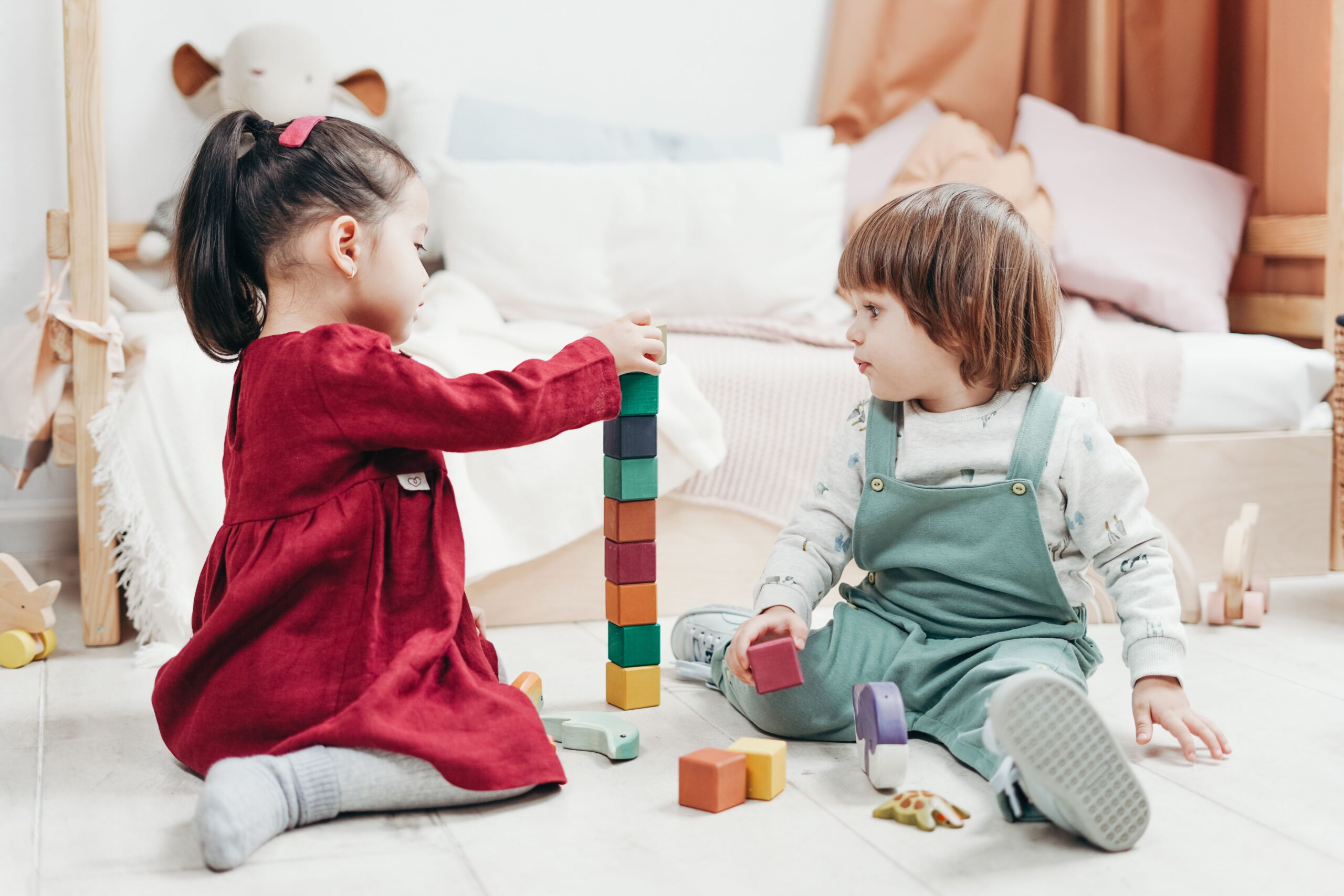 Two young children sit on the floor stacking colorful wooden blocks together in a cozy, softly lit room with a bed and stuffed animals in the background.