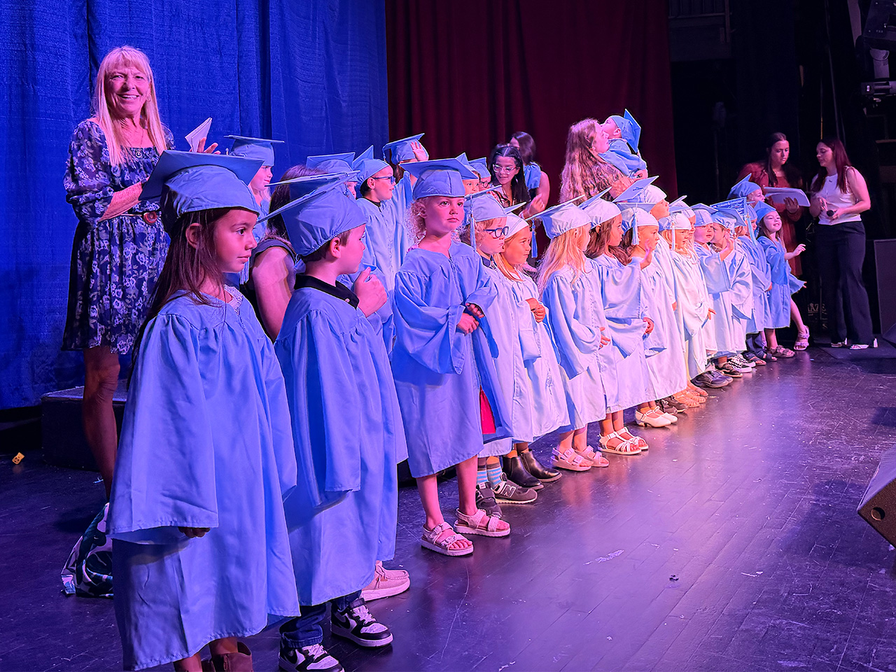 A group of young children in blue graduation gowns and caps stand in a row on a stage, smiling. Several adults stand behind them, and a woman on the left is clapping. Blue curtains form the background.