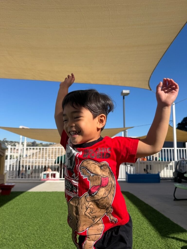 A young child wearing a red T-shirt with a dinosaur graphic joyfully raises both arms while playing outdoors on artificial grass under sunshades, with a playground and blue sky in the background.