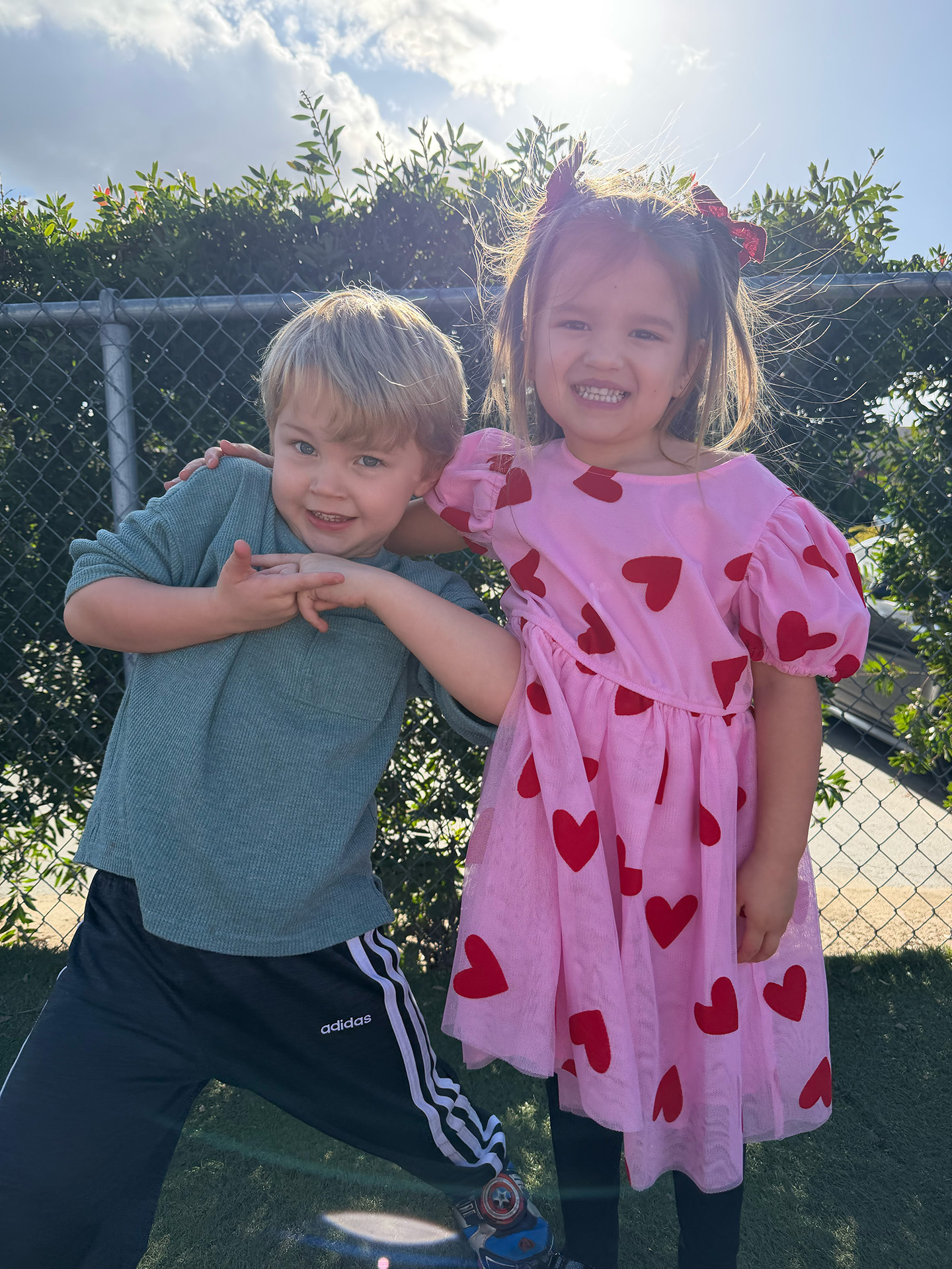 Two young children smile and pose playfully outdoors. The boy wears a green shirt and dark pants, while the girl wears a pink dress with red hearts. They stand in front of a fence with greenery, sunlight shining behind them.