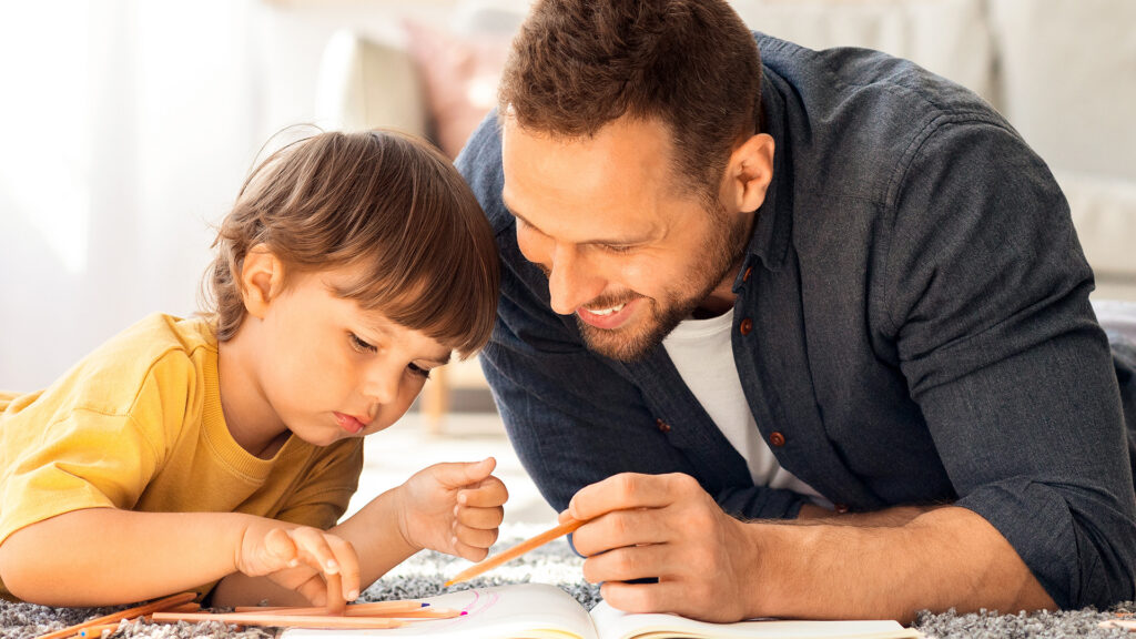 A man and a young child lie on the floor, smiling and drawing together on paper with colored pencils in a bright, cozy room.