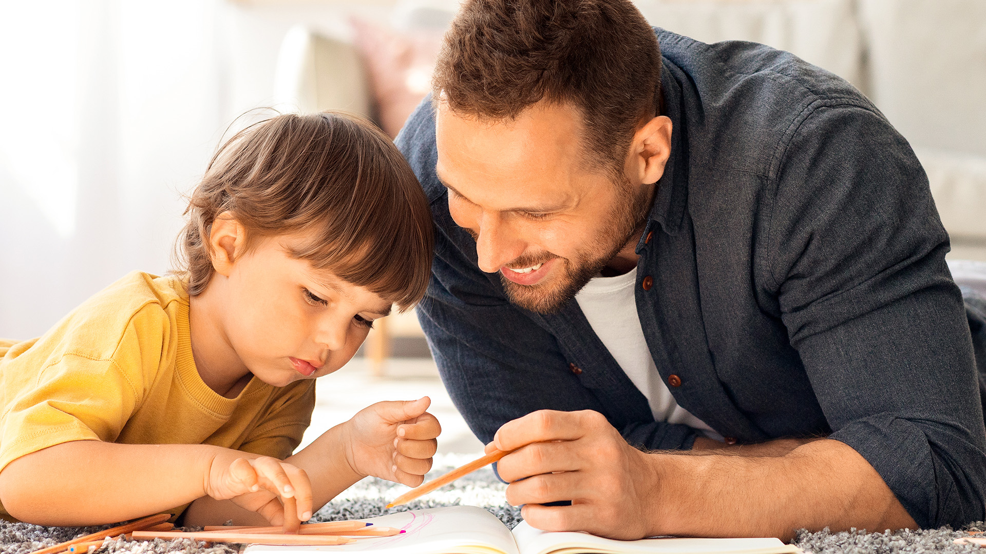 A man and a young child lie on the floor, smiling and drawing together on paper with colored pencils in a bright, cozy room.