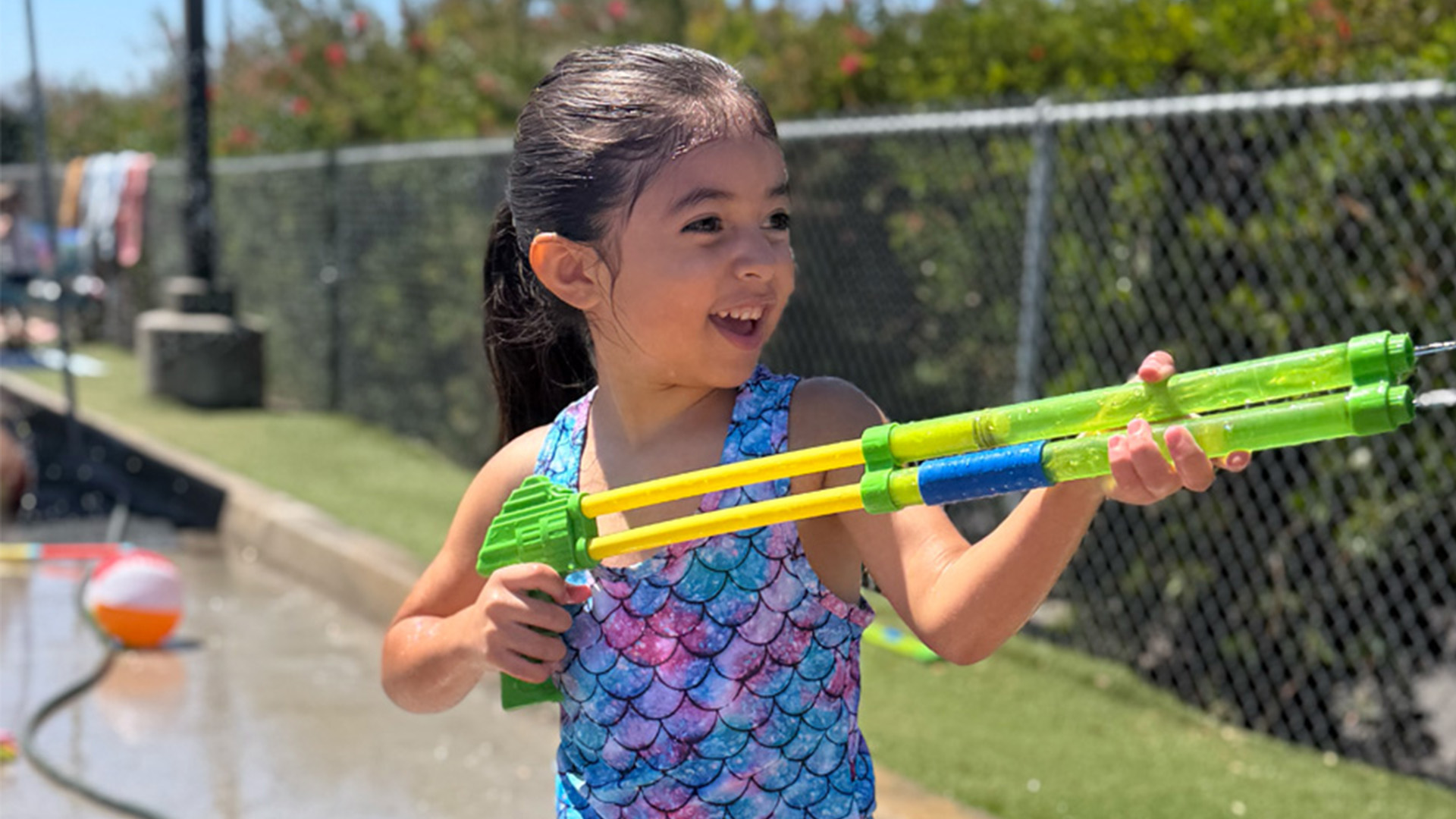 A young girl in a blue mermaid swimsuit smiles and aims a green water gun outdoors on a sunny day, with a fence, grass, and beach ball visible in the background—perfect inspiration for fun toddler activities and everyday learning through play.