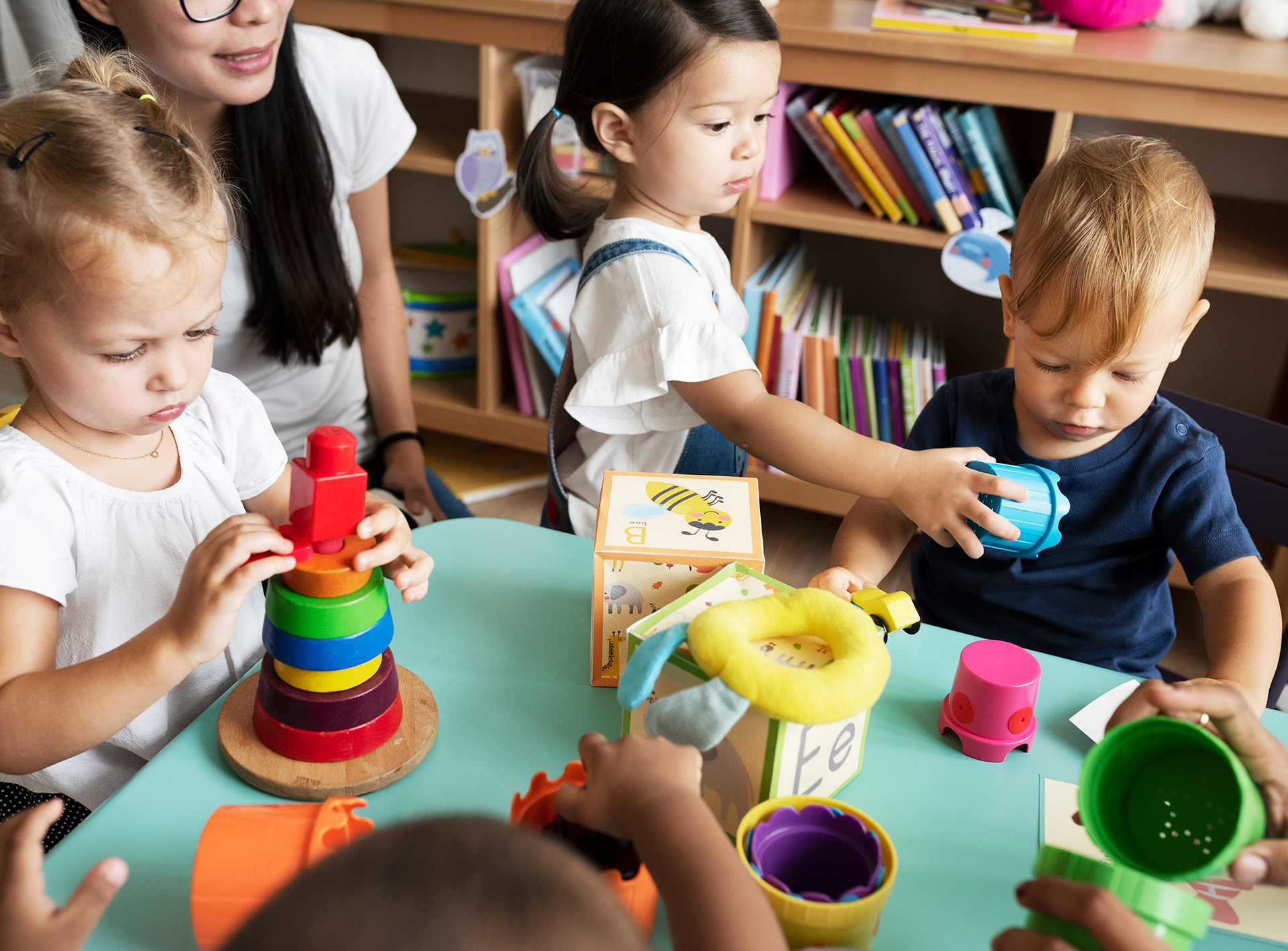 Four young children play with colorful educational toys at a round table in a Mission Valley classroom, with bookshelves in the background and an adult partially visible, supervising this engaging preschool activity.