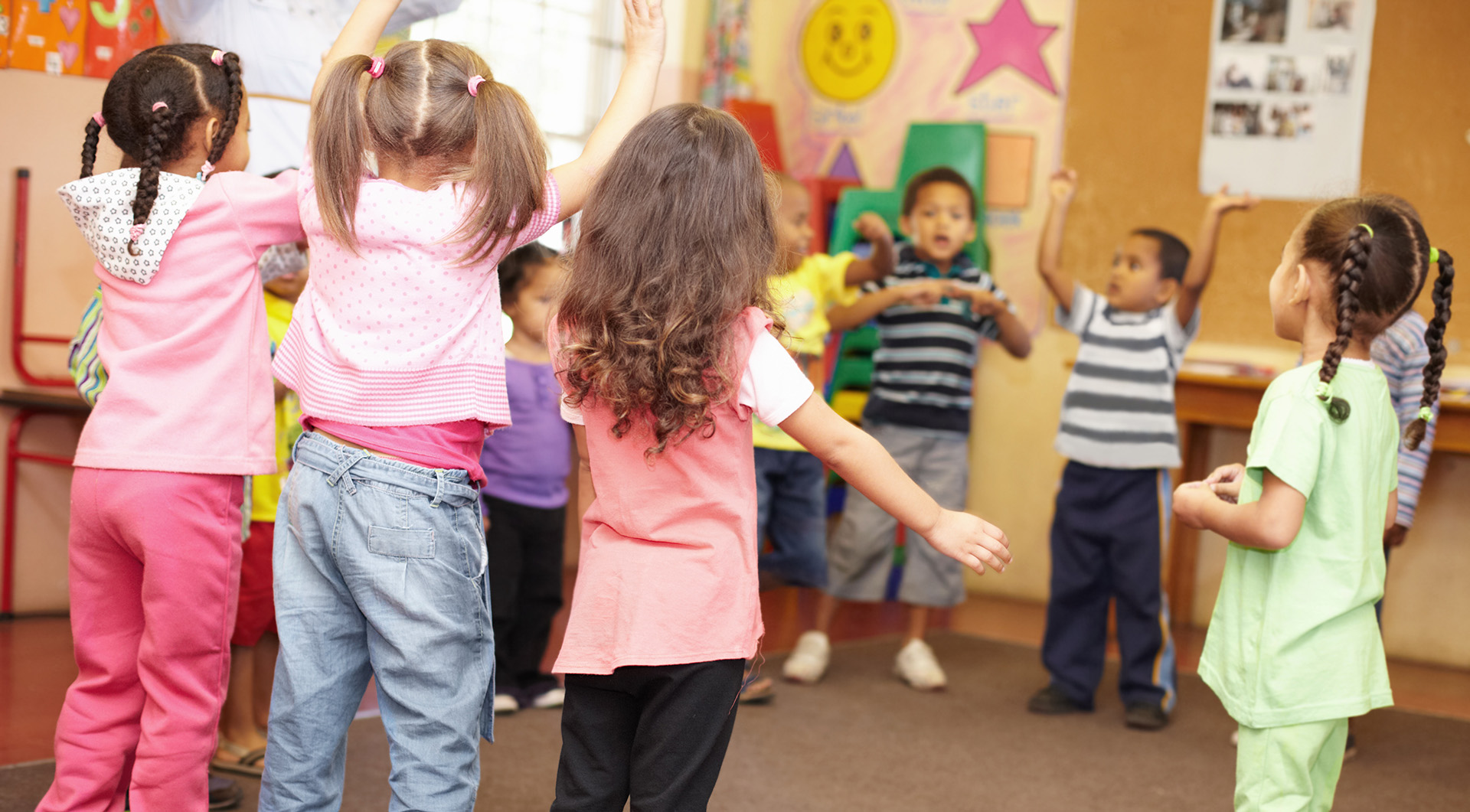 A group of young children stand in a classroom, raising their arms and playing together during a lively preschool tour. Colorful decorations and drawings adorn the walls in the background, making it easy to see why this San Diego classroom is sure to be a highlight on any parent’s checklist.